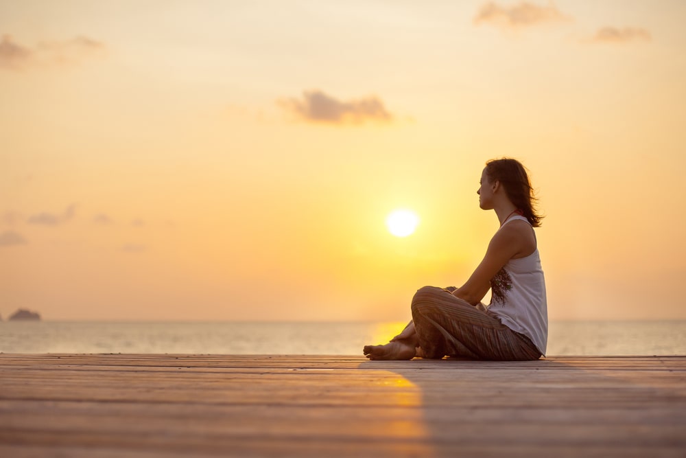 Person meditating on a beach, with the first rays of sunrise illuminating the surroundings-min
